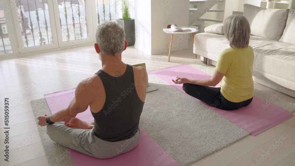 Fit serene retired older mid aged senior couple meditating learning virtual yoga class, doing breathing online exercise in living room at home using laptop. Digital fitness training for mental health