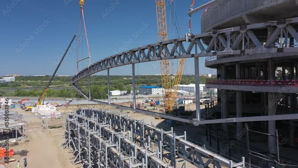 Lifting part of roof carcass with tower crane at sports stadium building construction site on riverbank aerial view Lifting part of roof carcass with tower crane at sports stadium building construction site on riverbank aerial view