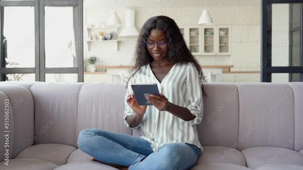 Young happy black African woman wearing glasses holding digital tablet sitting on couch at home learning online, surfing internet, doing e shopping in eshop using technology device relaxing on sofa.