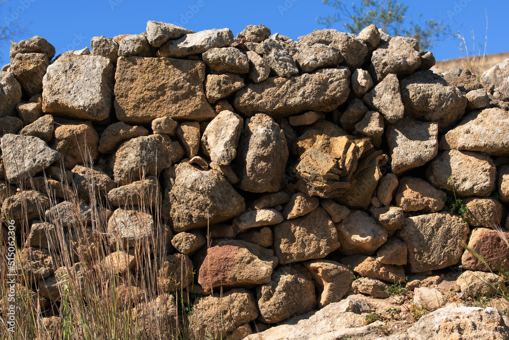 An ancient wall of rough stones stacked on top of each other in the traditional way in Sicily. Above is blue sky. There is dry grass in front of the wall. An ancient wall of rough stones stacked on top of each other in the traditional way in Sicily. Above is blue sky. There is dry grass in front of the wall.