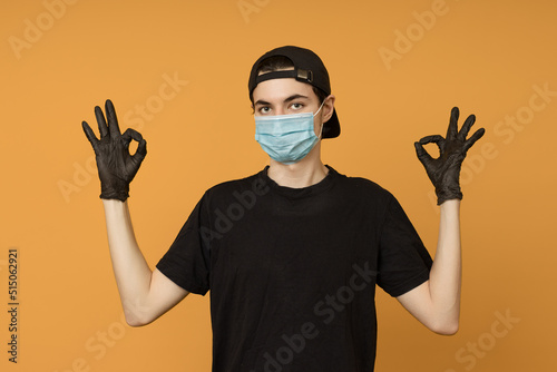 a young man in a black T-shirt, mask and gloves shows an OK sign with two hands, colored background