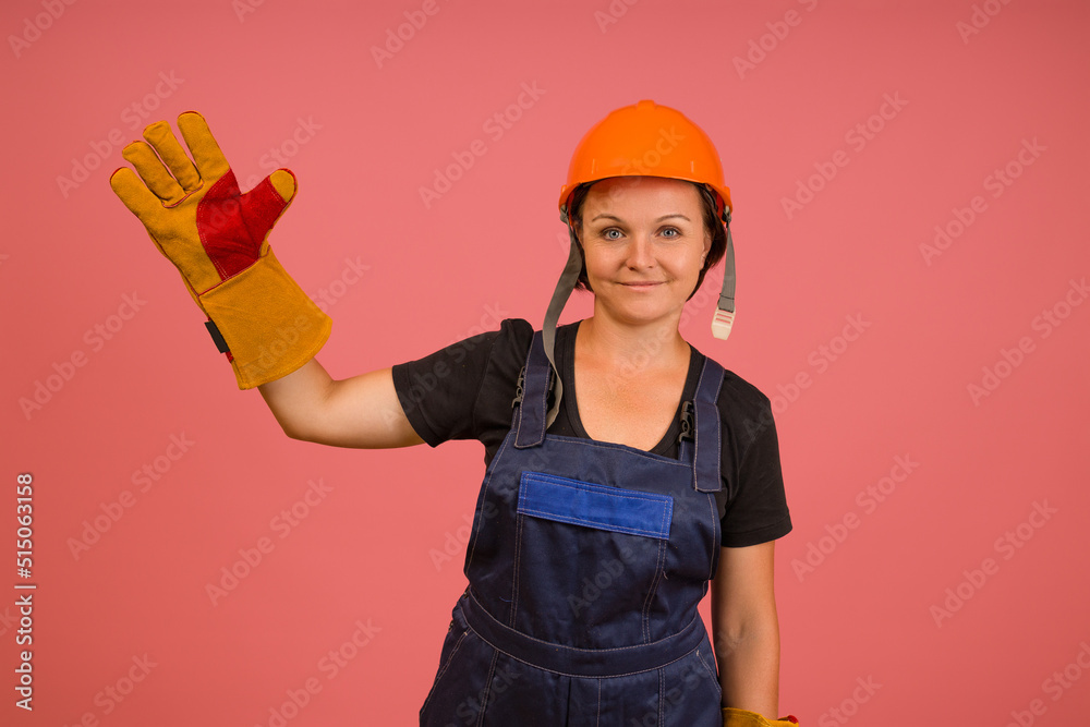 a woman in protective overalls, gloves and a helmet raised one hand up on a pink background