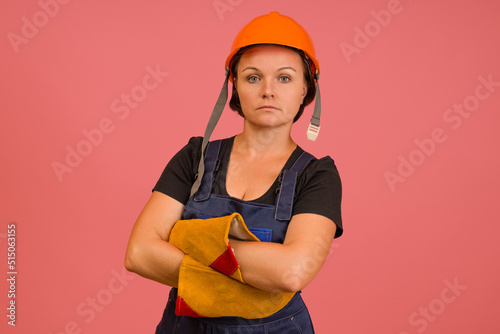 strong woman in protective overalls, gloves and a helmet crossed her arms over her chest on a pink background © Sergey