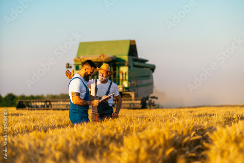 Canvas Print Grandfather and grandson, satisfied with the harvest, walk towards each other in the wheat field, rejoicing in their mutual success, while in the background the harvester harvests