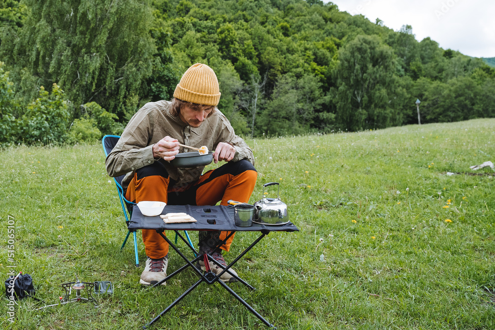 Breakfast in nature in the background of the forest, the guy eats fried eggs from the pan, portable folding table, kettle with tea, gas burner, male tourist eats Breakfast in nature in the background of the forest, the guy eats fried eggs from the pan, portable folding table, kettle with tea, gas burner, male tourist eats