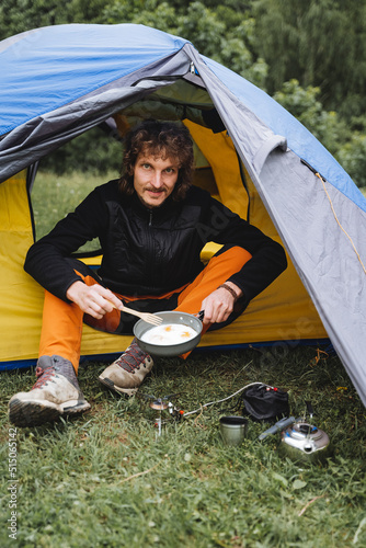 A guy sits in a tent holding a frying pan with scrambled eggs, a man has breakfast in nature at a campsite, cooking a meal on a hike on a gas burner.