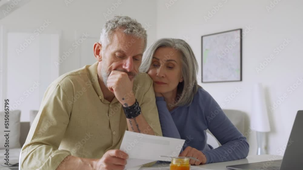 Older mature couple checking bank documents using laptop at home. Senior mid age retired man and woman reading paper bills, calculating pension or taxes, planning retirement finances, doing paperwork.