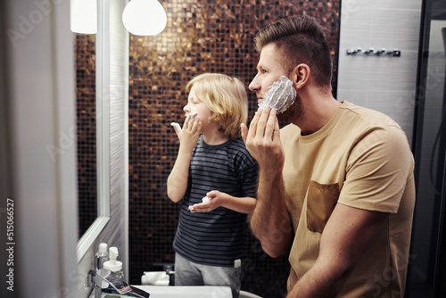 Side view portrait of father and son shaving together and looking in mirror in home bathroom