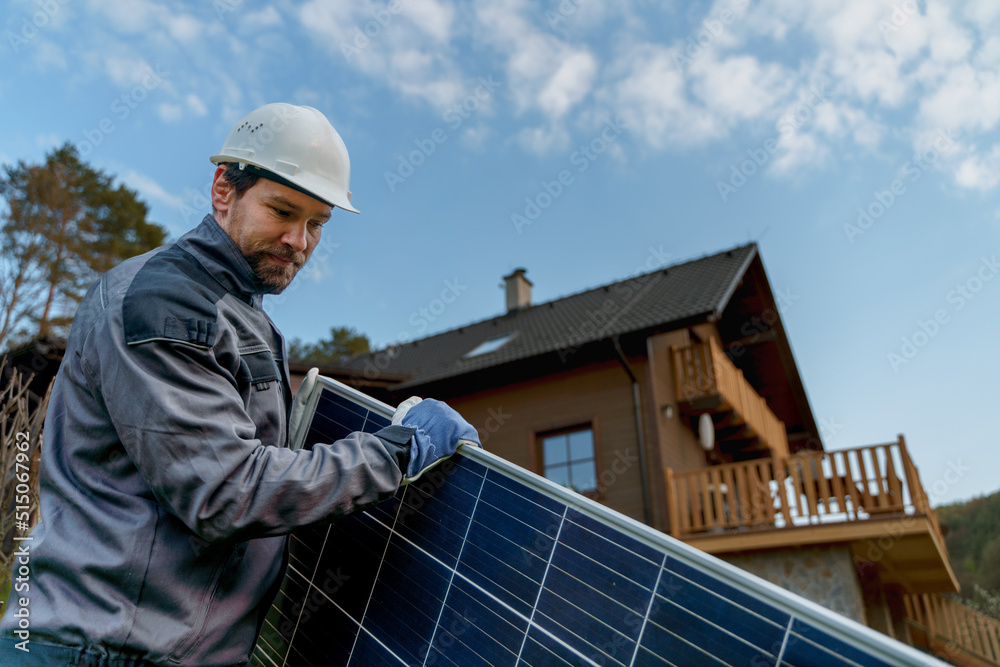 Smiling handyman solar installer carrying solar module while installing solar panel system on house.