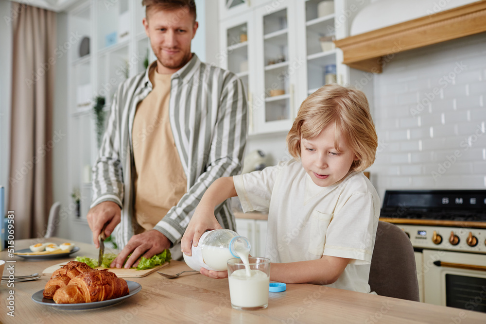 Warm toned portrait of cute blonde boy pouring glass of milk in cozy kitchen with father in background