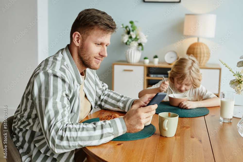 Side view portrait of father using smartphone at dining table while enjoying breakfast with son