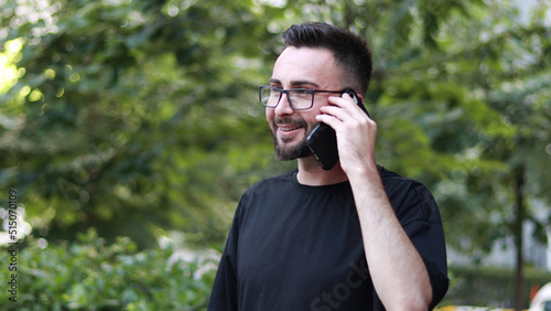 Portrait of young handsome man talking on the phone with beard and glasses. Smiling young man in black shirt. Outdoor nature background