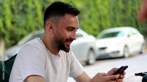 Bearded and handsome young man using mobile phone outdoors. Happy smiling man in white t-shirt looking at mobile phone screen and reading messages