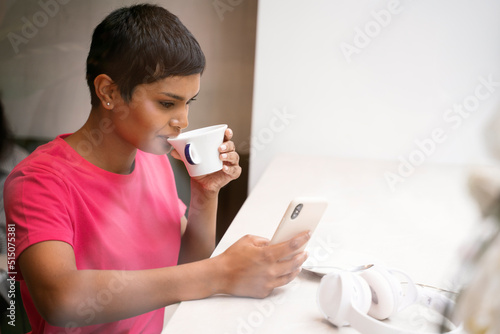 Beautiful Indian woman holding mobile phone reading text message, drinking coffee in cafe. Successful asian freelancer working online, check email sitting at workplace 