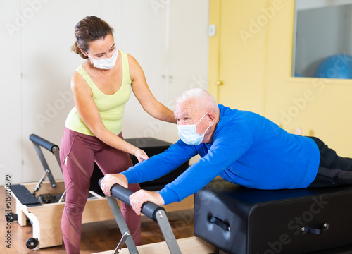 Focused senior man in protective face mask doing exercises on Pilates reformer under supervision of qualified female trainer. Concept of physical activity in pandemic © JackF