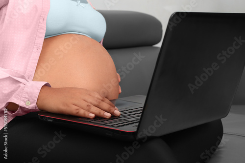 Latina pregnant woman using laptop on legs sitting on couch