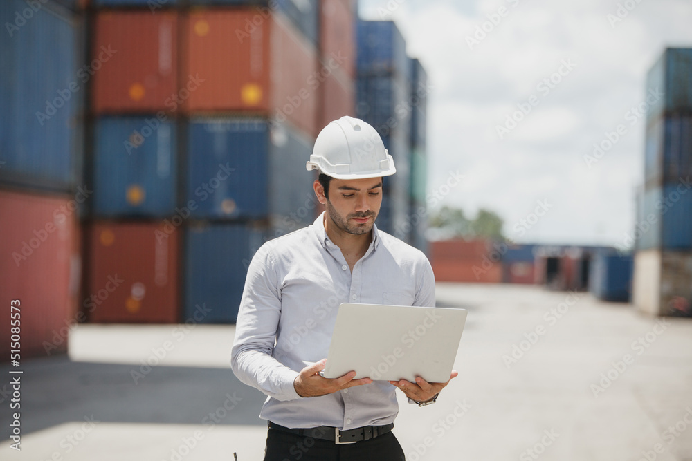 Engineer worker in causual suit standing in shipping container yard holding laptop with smile. Import and export product. Manufacturing transportation and global business concept Engineer worker in causual suit standing in shipping container yard holding laptop with smile. Import and export product. Manufacturing transportation and global business concept