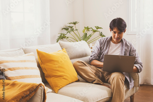 Handsome asian man t-shirt sits lobby sofa in the living room of his home connected to reading Internet messages. Look at your laptop computer screen cozy little corner of your home.