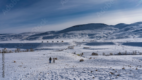 Fototapeta Naklejka Na Ścianę i Meble -  Two people walk through an endless snow-covered valley. Spherical glamping houses are visible in the distance and ahead. Mountains against a background of blue sky and clouds. Altai