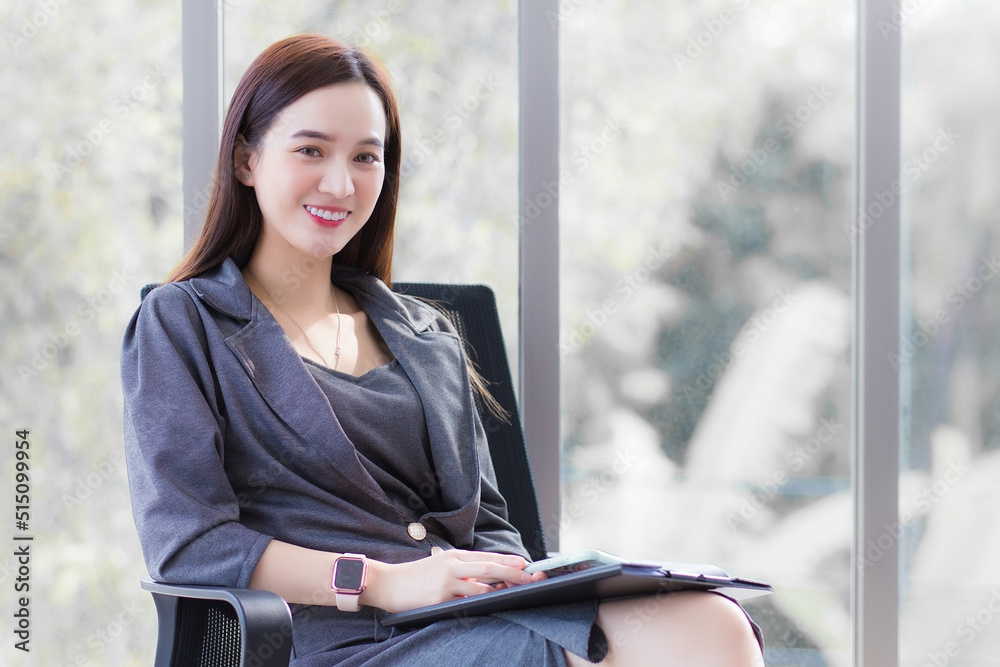 Professional Asian business woman in dark grey dress sits on chair in office room and works happily with a mirror as a background. Professional Asian business woman in dark grey dress sits on chair in office room and works happily with a mirror as a background.