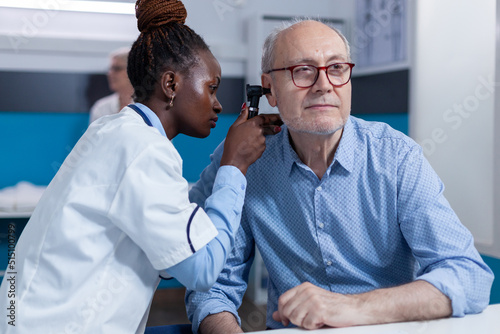 Clinic otology specialist consulting senior patient using otoscope to check ear infection. Hospital otologist examining sick retired man internal ear condition while in doctor cabinet. photo