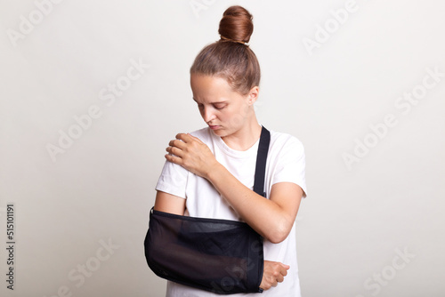 Indoor shot of unhealthy woman wearing casual T-shirt standing in arm sling and feeling pain in shoulder, touching painful part of the body, car accident, posing isolated on light gray background. photo