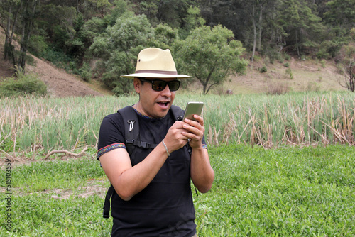 Latin adult man with hat, shorts and sunglasses uses his cell phone in the middle of the forest outdoors to make calls, receive messages, locate on the map because he is lost 