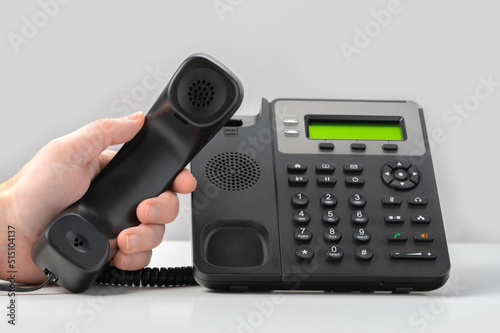 hand holding voip telephone receiver on gray background. black office landline voip phone with handset up on table. photo