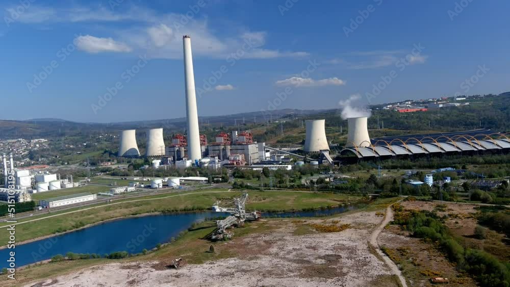 Thermal power plant with smoking chimney, lake in front with an old excavation machine, bright and sunny afternoon. Drone shooting traveling backwards slowly. As Pontes, Lugo, Spain Thermal power plant with smoking chimney, lake in front with an old excavation machine, bright and sunny afternoon. Drone shooting traveling backwards slowly. As Pontes, Lugo, Spain