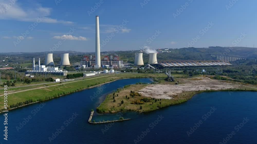 Thermal power plant with its smoking chimney, a lake in front with an old industrial machine, a bridge on the right, bright and sunny afternoon. Drone shot rising. As Pontes, Lugo, Spain Thermal power plant with its smoking chimney, a lake in front with an old industrial machine, a bridge on the right, bright and sunny afternoon. Drone shot rising. As Pontes, Lugo, Spain