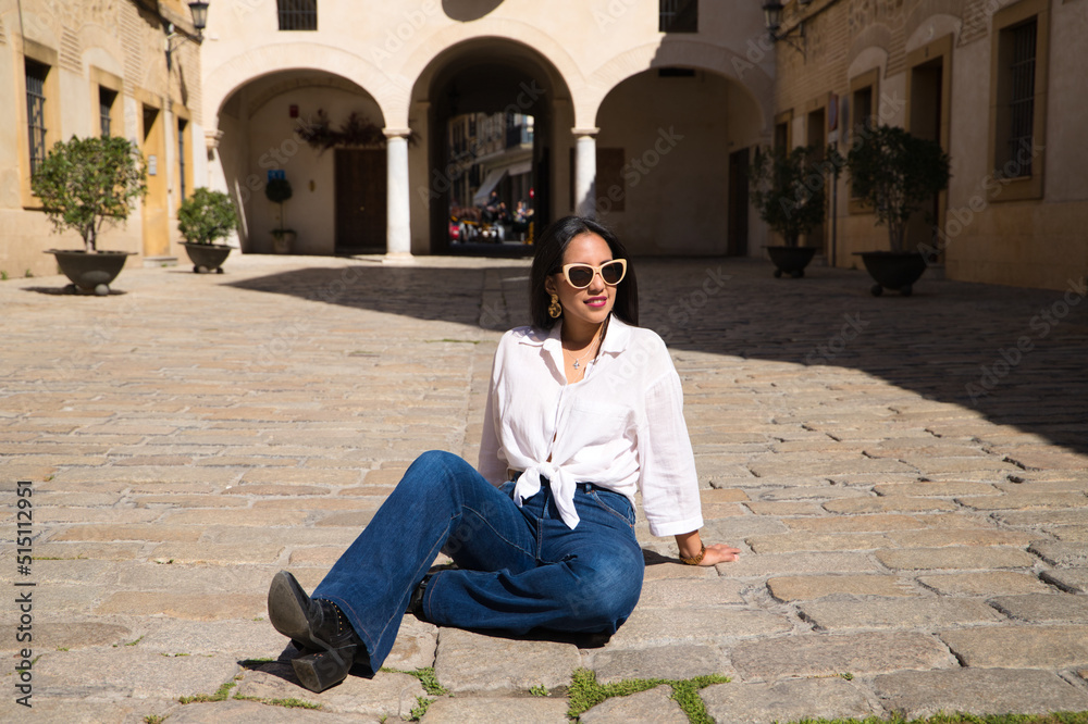 young, beautiful, brunette south american woman is sitting on the brick floor in a typical mediterranean city sunbathing. The woman poses for the photo. Holiday and travel concept