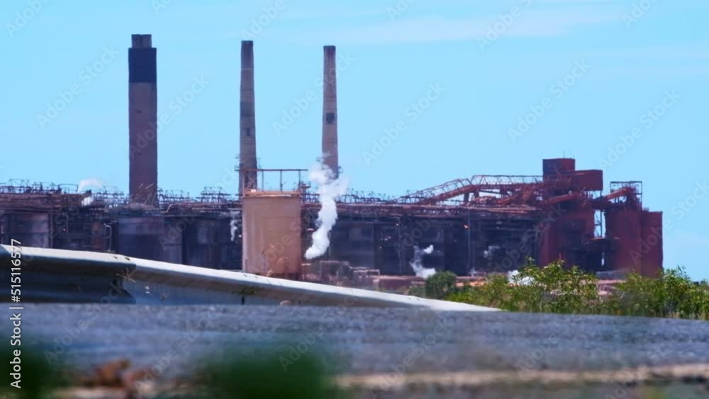 View of large industrial plant from the road with steam coming out and bright blue sky.