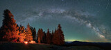 Panorama of starry sky, Milky Way over mountain hills. Silhouettes of two travelers with tent who warming by bonfire. Forest, tops of mountains with light from settlements in the distance on backdrop.