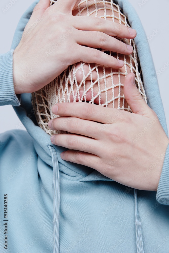 a close vertical, conceptual studio photograph of a strange, tortured man in a light blue hoodie covering his face with his hands, standing on a light background with a hood on his head