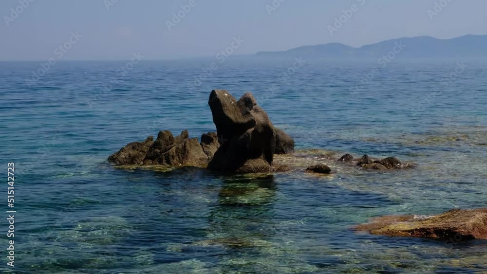 Zoom out to colorful rocks in the shallow Mediterranean water in the middle of a sunny day. Rocky beach in Assos, Canakkale at Aegean cost of Turkey. Lesbos Island in the background. Zoom out. Zoom out to colorful rocks in the shallow Mediterranean water in the middle of a sunny day. Rocky beach in Assos, Canakkale at Aegean cost of Turkey. Lesbos Island in the background. Zoom out.