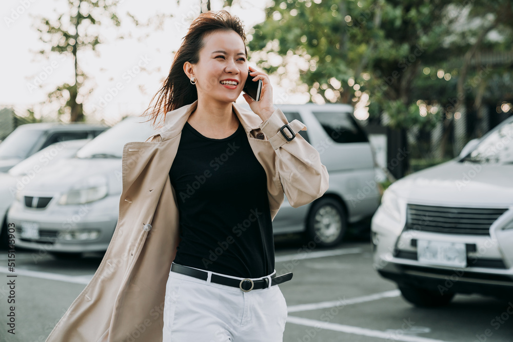 successful asian woman manager in smart casual is walking by a city car parking lot while having a pleasant business talk on the smartphone on a windy day.