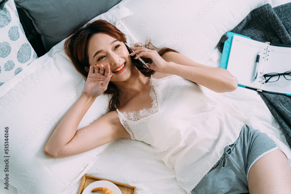 top angle shot cheerful asian girl lying in the bedroom is touching her face with a smile while chatting on the phone in a carefree morning at home. top angle shot cheerful asian girl lying in the bedroom is touching her face with a smile while chatting on the phone in a carefree morning at home.