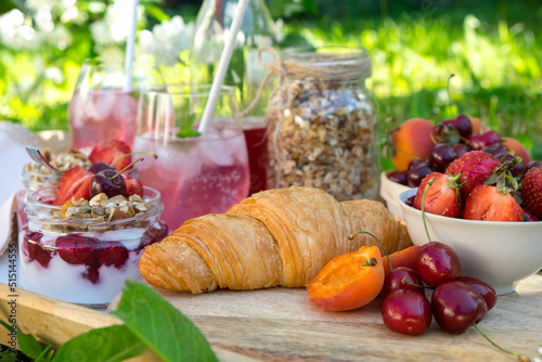 Healthy breakfast of croissant, fruits and berries and parfe with yogurt and granola served on a tray in the garden with green grass and white flowers in the background. 