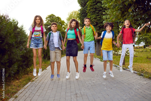 Funny elementary school students with backpacks on their shoulders have fun jumping together on path in park. Cheerful preteen boys and girls in casual clothes stand in row holding hands and jumping.