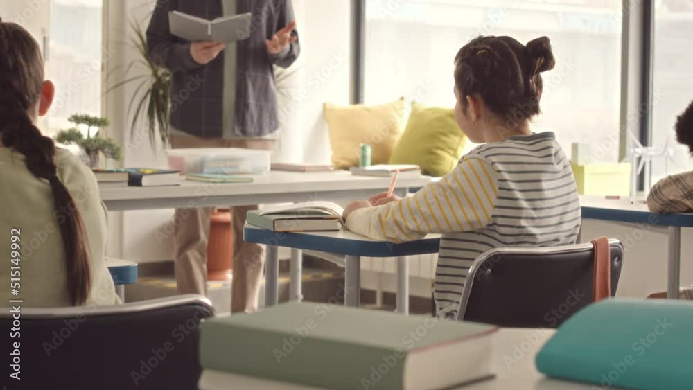 Waist up slowmo of elementary school girls using cheat sheets during lesson in classroom while male teacher standing in front of them with book in hands Waist up slowmo of elementary school girls using cheat sheets during lesson in classroom while male teacher standing in front of them with book in hands