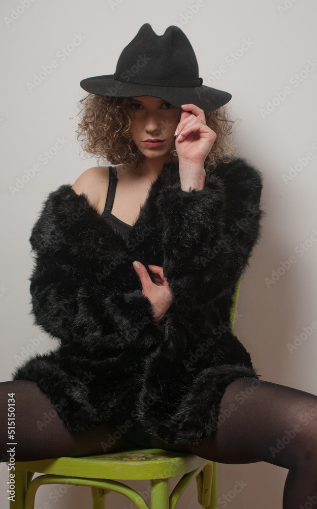 Portrait of a fashionable model with black fur sitting in a green chair in studio. young beautiful woman in black pantyhose .Portrait of attractive woman sitting in chair with black hat Portrait of a fashionable model with black fur sitting in a green chair in studio. young beautiful woman in black pantyhose .Portrait of attractive woman sitting in chair with black hat