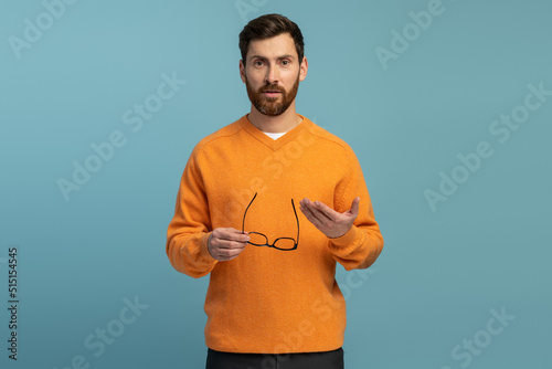 Waist-up shot of serious handsome man with beard taking off glasses and holding eyewear in hand with misunderstanding look gazing at camera