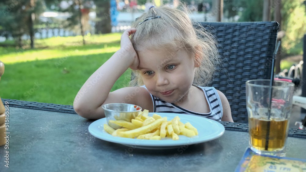 Little girl eat french fries. Close-up of blonde girl takes potato chips with her hands and tries them sitting in street cafe on the park. Little girl eat french fries. Close-up of blonde girl takes potato chips with her hands and tries them sitting in street cafe on the park.