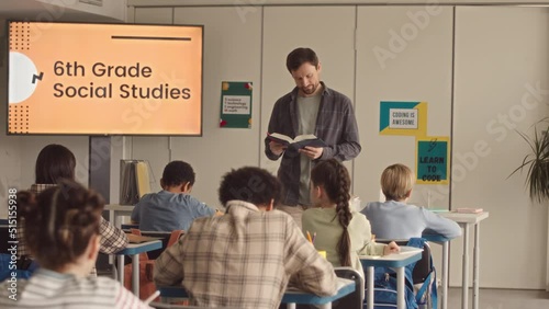 Medium slowmo of mid adult male school teacher of Social Studies reading topic from student book to children taking notes in copybooks while listening photo