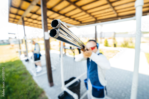 Canvas Print Blurred background with focus on front of shotgun barrel of adult man in sunglasses, protective headphones and a rifle vest practicing fire weapon shooting