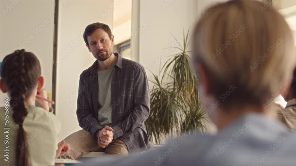Slowmo of kind mid adult male teacher talking to group of middle school students while sitting on desk in modern classroom during lesson Slowmo of kind mid adult male teacher talking to group of middle school students while sitting on desk in modern classroom during lesson