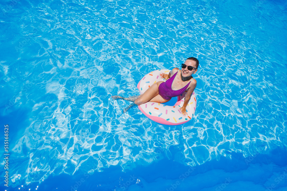Beautiful slim young woman in a swimsuit enjoying the water park floating in an inflatable big ring on a sparkling blue pool smiling at the camera. Summer vacation.
