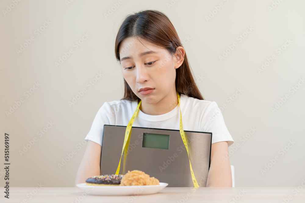 Diet, attractive asian young woman, girl restrained to eat doughnut, bakery and fried chicken, fast food to lose, loss weight, hugging weight scales on table at home. Passion, temptation when hungry.