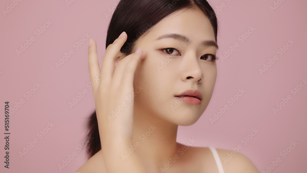 Close up face of beautiful young Asian woman with clear skin lifting eyes with finger on pink background.