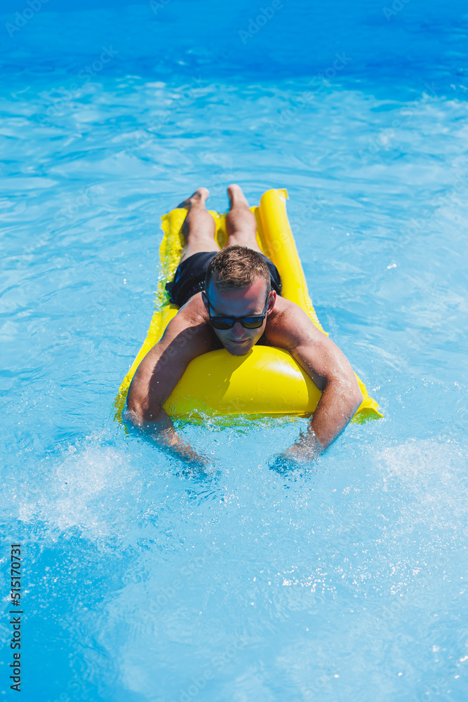 Attractive young man in sunglasses and shorts is relaxing on an inflatable yellow mattress in the pool. Summer vacation